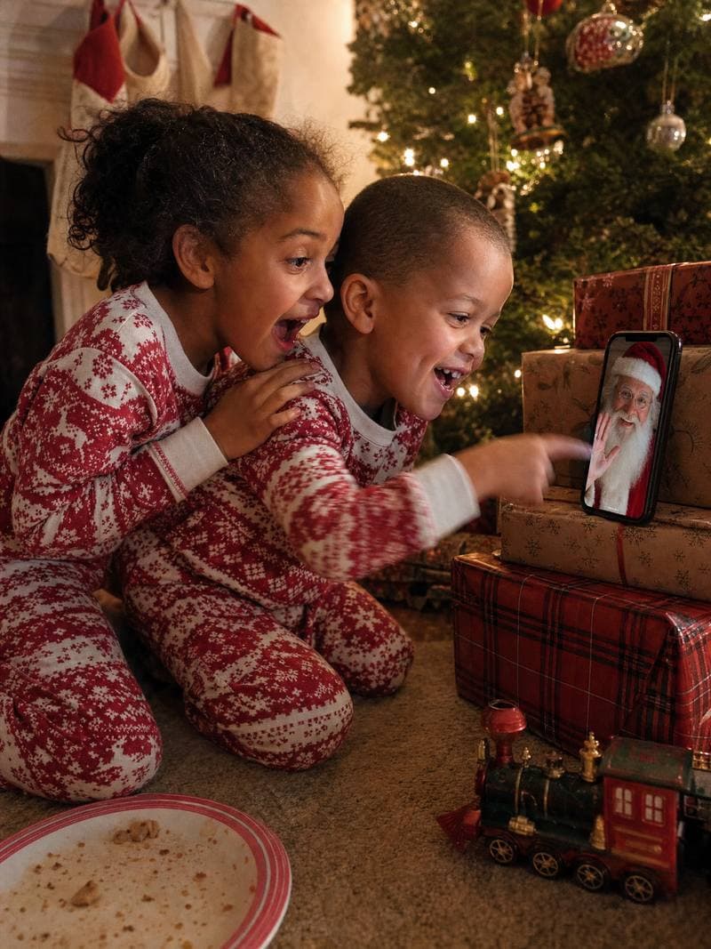 Brother and sister in pyjamas pointing at Santa on a phone by the tree